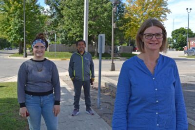 Three people stand socially distanced on a shady sidewalk on a city street