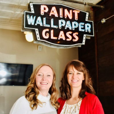 Kayla and Valerie stand underneath a vintage neon sign advertising paint, wallpaper and glass