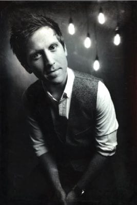 A black and white image shows Josh in a shirt and vest in front of lights suspended by strings.