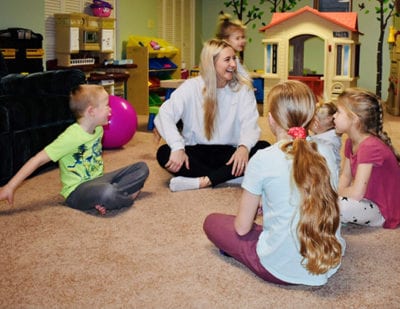 Angela sits with a group of kids on a carpeted floor playing duck, duck, gray duck