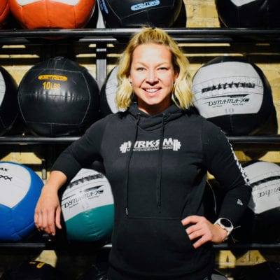 Heather stands in front of racks of large medicine balls and holds one under the crook of her arm.