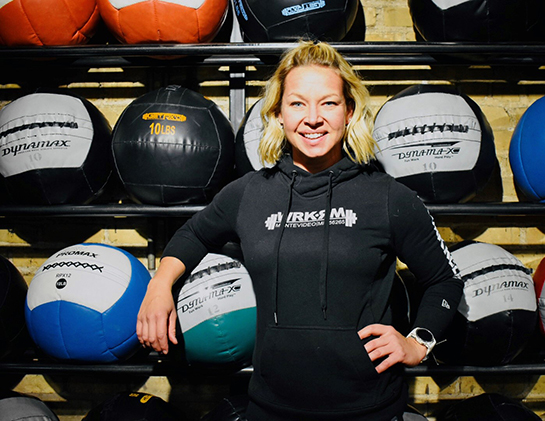 Heather stands in front of racks holding colorful medicine balls, with a teal one tucked under her arm