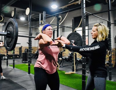 Heather guides a member's elbows as the person lifts a bar with weights