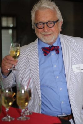 Greg holds up a glass for a toast and wears a white suit coat over a blue shirt with a black and red striped bowtie