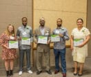 The entrepreneurs graduating from Elevate Business Academy stand shoulder-to-shoulder holding framed certificates.