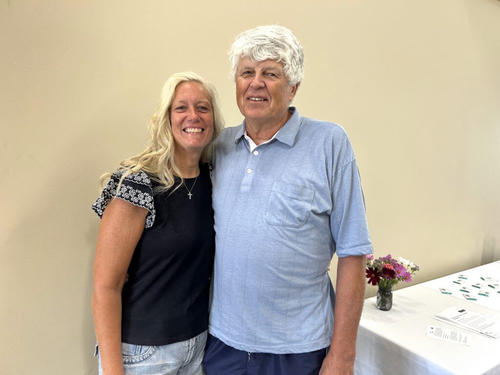 Two adults stand close together and smile at the camera. There is a table with small bouquets of flowers and papers in the background against a beige wall.