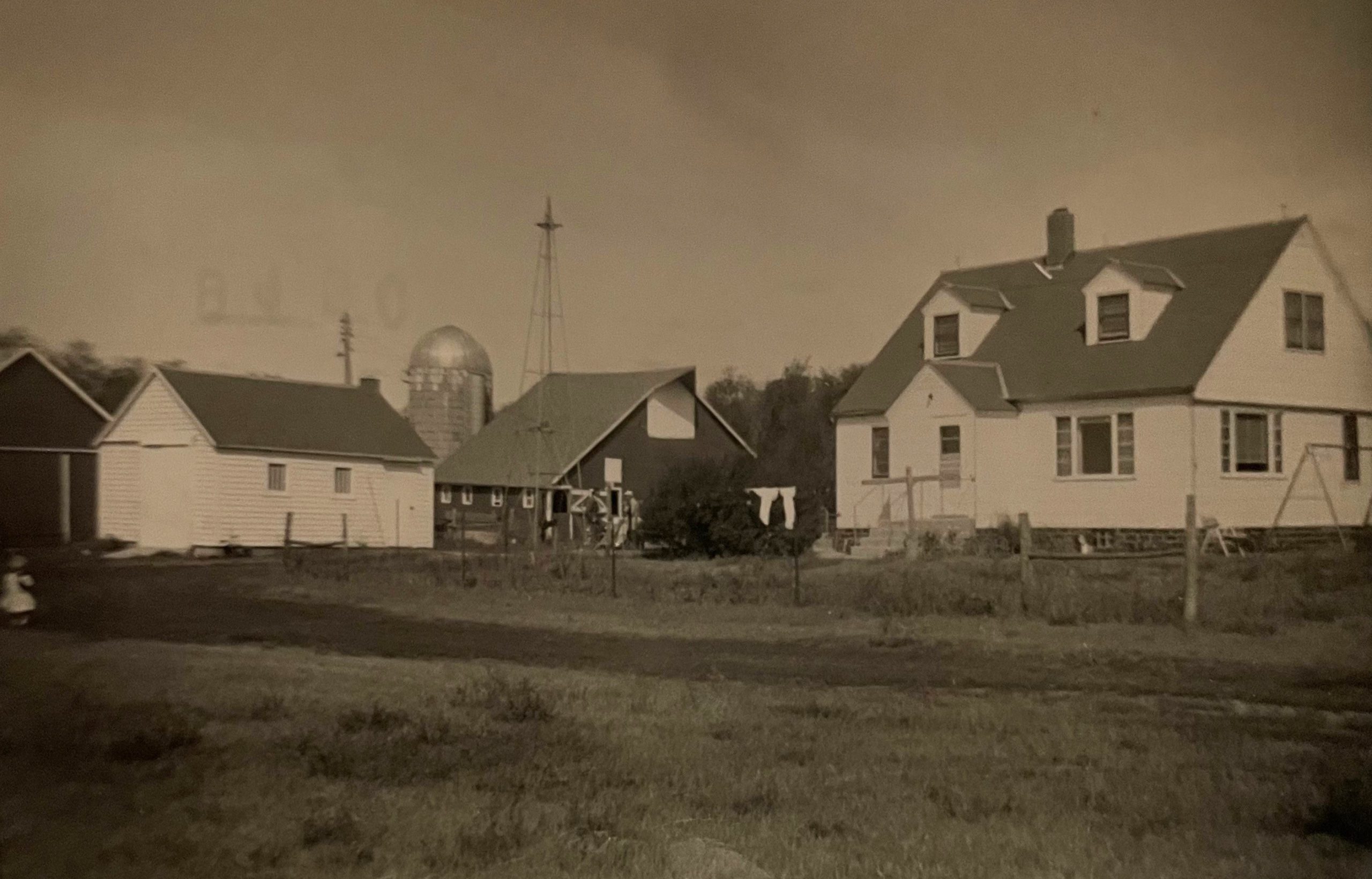 A black and white photo of a rural farm with a two-story house, a barn, a silo, a windmill, and a clothesline with laundry hanging between buildings; grassy yard and fenced area in the foreground.