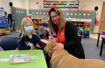 Marie sits at a table in a classroom with a student and her therapy dog, Ally