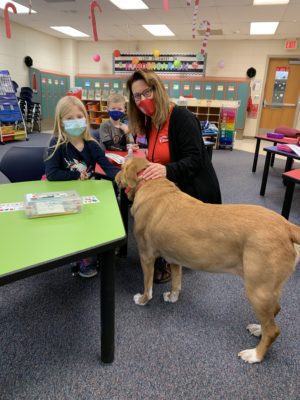 Marie sits in a classroom at a small table with a young student and support dog Ally