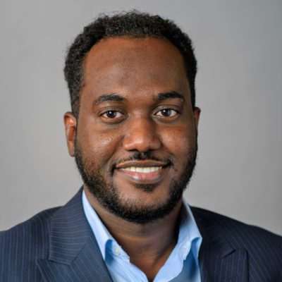 Khalif is pictured in a blazer and button-up shirt in front of a gray background