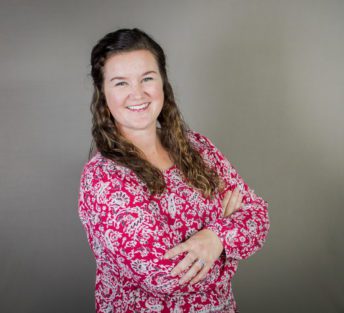 Meghan poses for a formal head shot in front of a gray background wearing a red paisley top