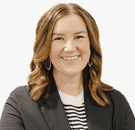 A woman with wavy light brown hair, wearing a black blazer over a black-and-white striped top, smiling at the camera against a plain white background.