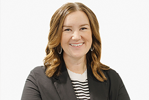 A woman with wavy light brown hair, wearing a black blazer over a black-and-white striped top, smiling at the camera against a plain white background.