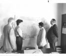 A black and white photo shows four people gathered around a map of the U.S. on a wall