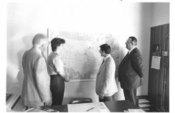 A black and white photo shows four people gathered around a map of the U.S. on a wall