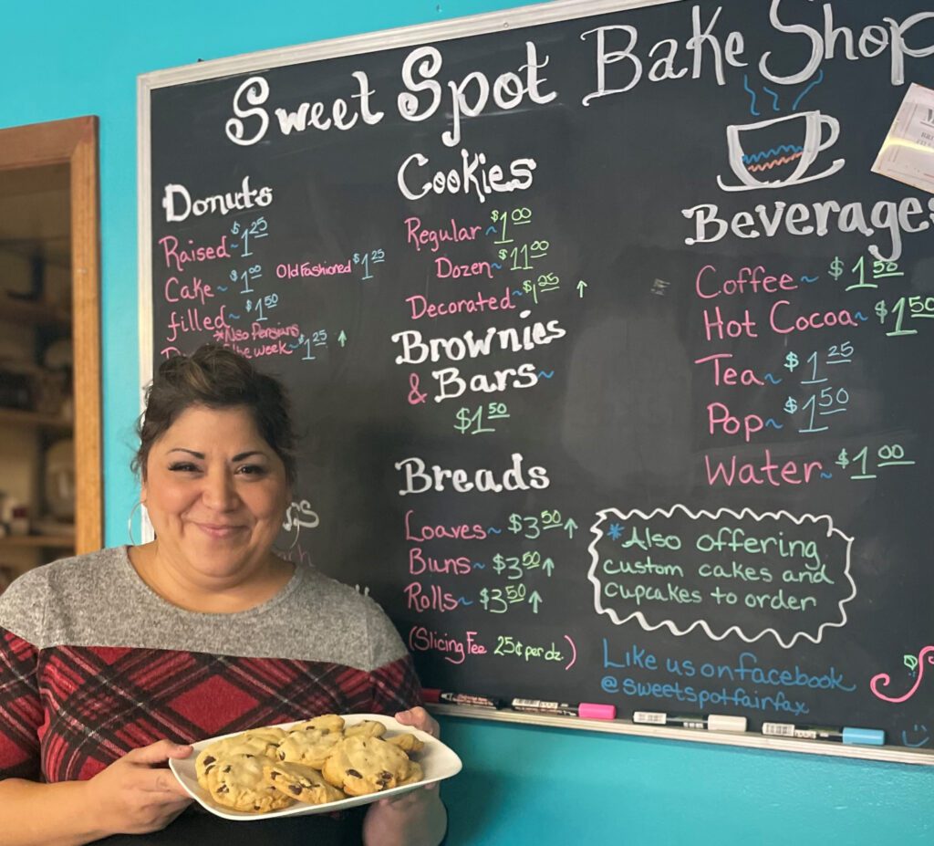 Tina holding a plate of cookies in front of a chalkboard sign listing bakery items