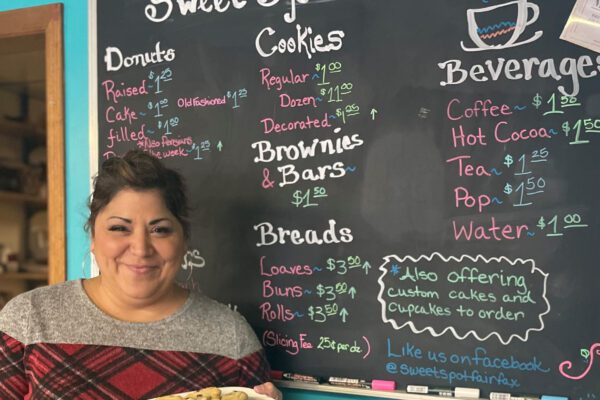 Tina holds a plate of cookies and stands in front of a chalkboard sign listing bakery items