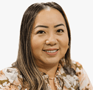 A woman with long, straight brown hair and dark eyebrows smiles at the camera. She is wearing a floral blouse and is posed against a plain, white background.