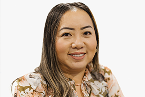 A woman with long, straight brown hair and dark eyebrows smiles at the camera. She is wearing a floral blouse and is posed against a plain, white background.