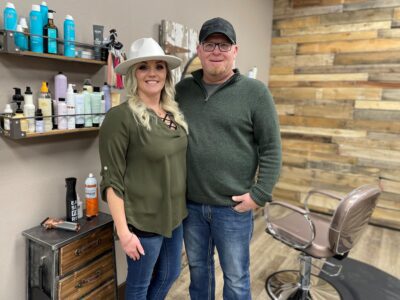 Chauntel and Mark Renne stand inside Rustic Beauty Salon with shelves of hair care products in the background and a salon chair next to them.