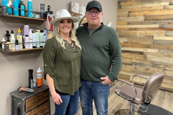 Chauntel and Mark Renne stand inside Rustic Beauty Salon with shelves of hair care products in the background and a salon chair next to them.