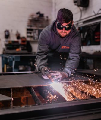 Mark wears welding goggles as he works on a piece of metal, sending sparks flying with a welding torch.