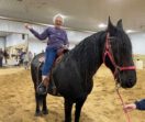 An older adult smiles as she sits in the saddle on a black horse in an indoor riding arena.