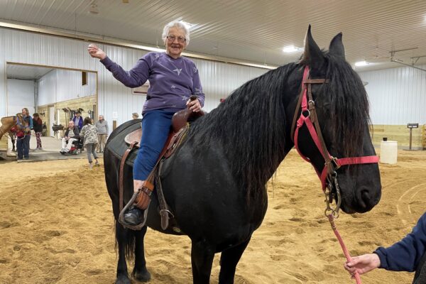 An older adult smiles as she sits in the saddle on a black horse in an indoor riding arena.