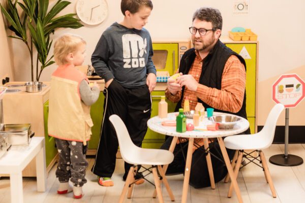 A grown-up and two kids play together around a child-size table covered with toy kitchen items.