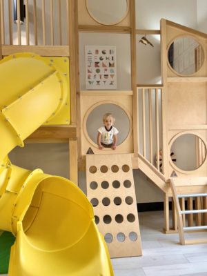 A child peeks out from a circle that's part of a large indoor play structure with a yellow slide.