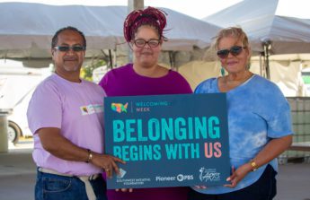 A group of three people gather at the Worthington Welcoming Week celebration and hold up a sign that reads Belonging Begins with US