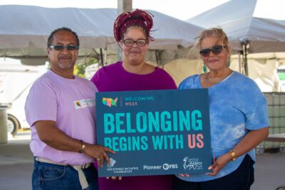 A group of three people gather at the Worthington Welcoming Week celebration and hold up a sign that reads Belonging Begins with US