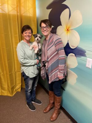 Skie holds therapy dog Koda and stands next to Jill near a wall mural of large white flowers.