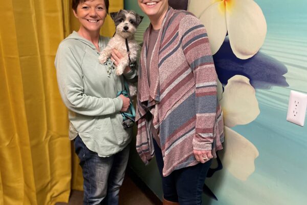 Skie holds therapy dog Koda and stands next to Jill near a wall mural of large white flowers.