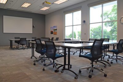 A view of a conference room that has large south-facing windows, a white screen on the far wall and moveable tables and chairs