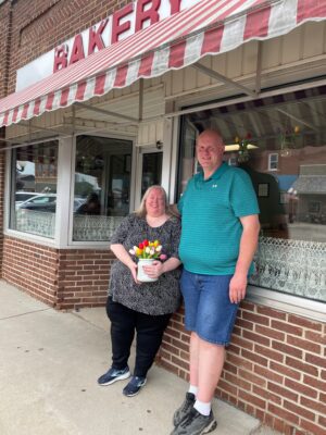 Carl and Stacie stand outside the bakery's storefront under a red and white striped awning.