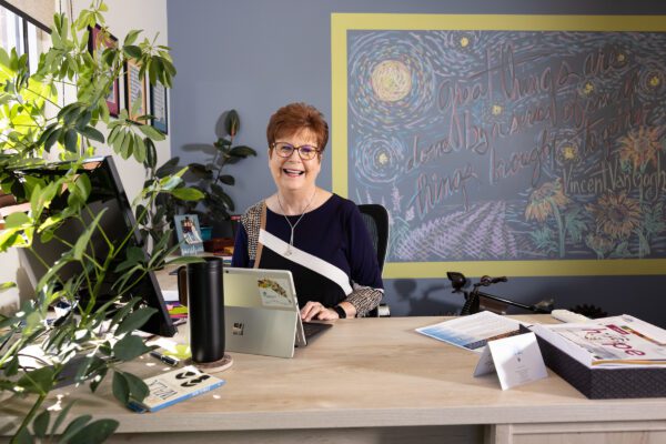 Diana sits at her laptop in her office with a large plant and a wall mural in the background.