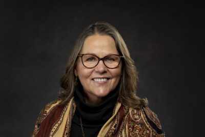 Kris poses for a formal head shot wearing a patterned caftan, black blouse and glasses.