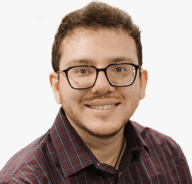 A person with short curly hair, glasses, and a trimmed beard smiles at the camera. They are wearing a maroon and blue checkered shirt against a plain white background.