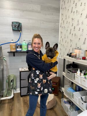 Kelly inside her dog grooming business holding a small dog that's wearing a sweatshirt.