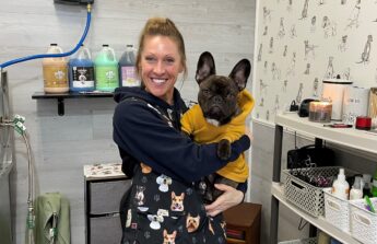 Kelly stands inside her dog grooming business holding a small dog that's wearing a yellow sweatshirt.