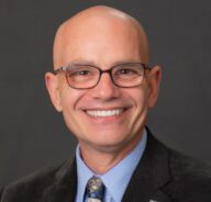 Terry poses for a formal head shot wearing a blazer, blue shirt, tie and glasses.