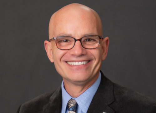 Terry poses for a formal head shot wearing a blazer, blue shirt, tie and glasses.