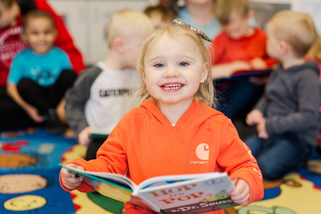 young girl at a child care center reading a children's book and smiling