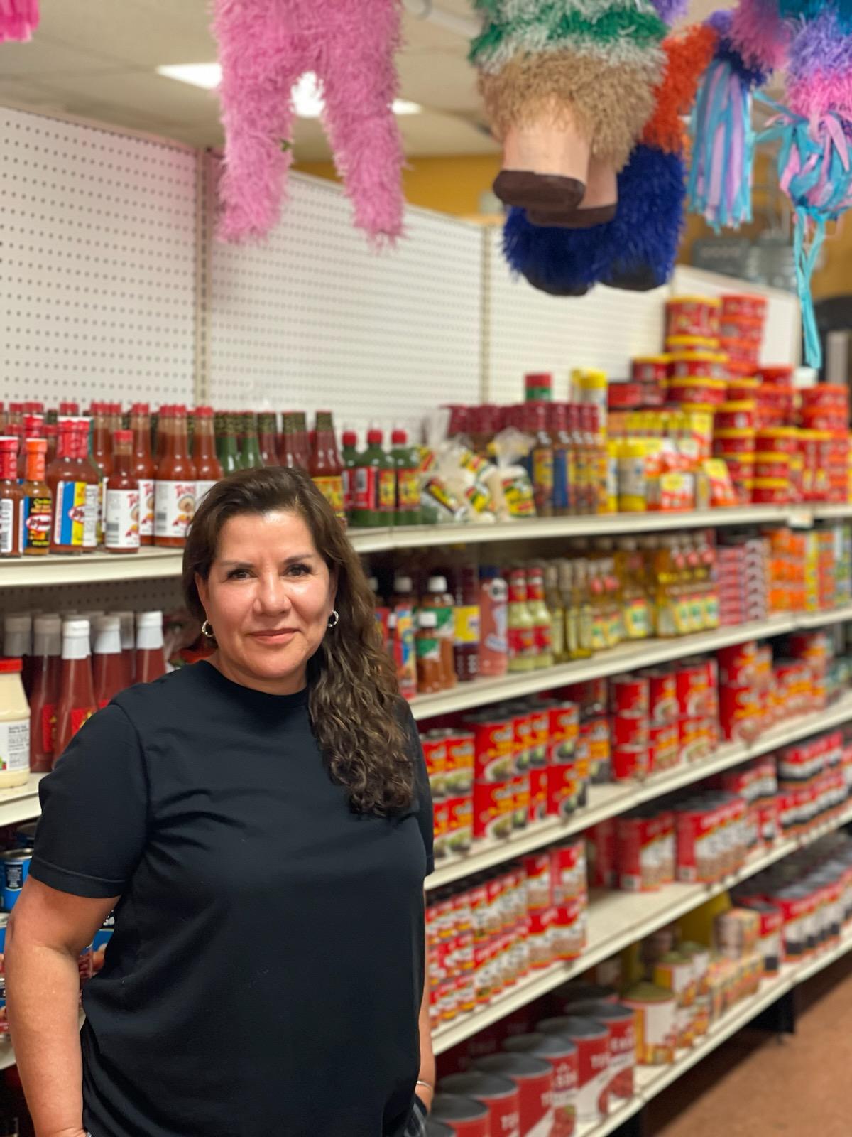 Maria Parga standing in a store with shelves of food behind her and pinatas hanging above her head.