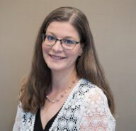 Erica poses for a formal head shot wearing glasses and a white lace cardigan over a black blouse.
