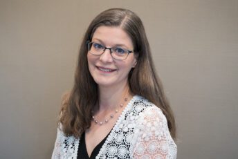 Erica poses for a formal head shot wearing glasses and a white lace cardigan over a black blouse.