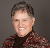 Theresa poses for a formal head shot wearing a brown mock turtleneck and patterned woven jacket.