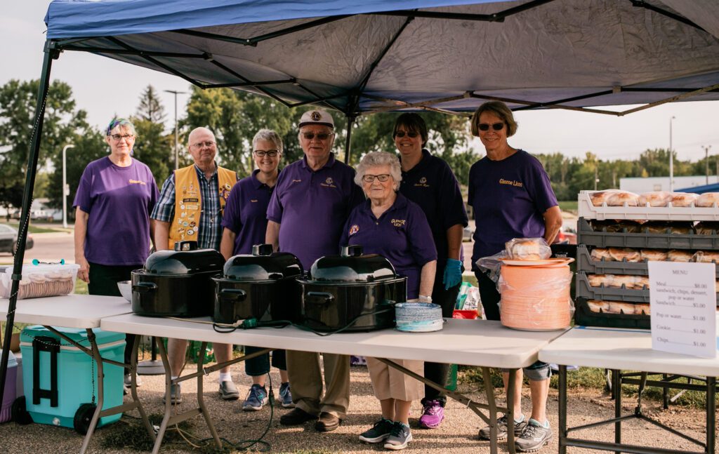 A group of volunteers wearing purple Lions Club T-shirts serves food at a community block party.