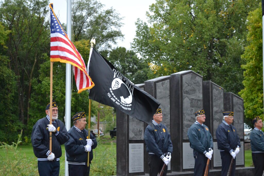 Two veterans hold up the United States flag and the POW MIA flag while other veterans stand at ease near the veterans memorial.